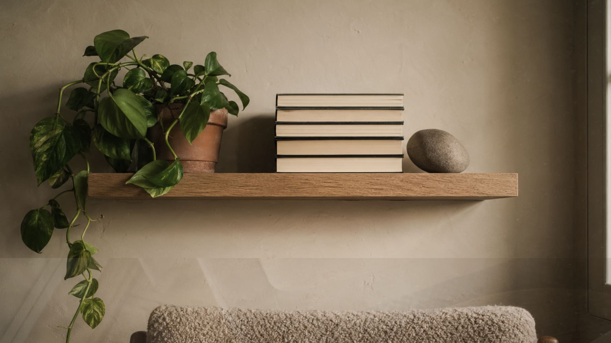 japandi reading nook floating oak shelf with books arranged pages-facing-out, trailing pothos in terracotta pot, and smooth river stone bookend on warm plaster wall
