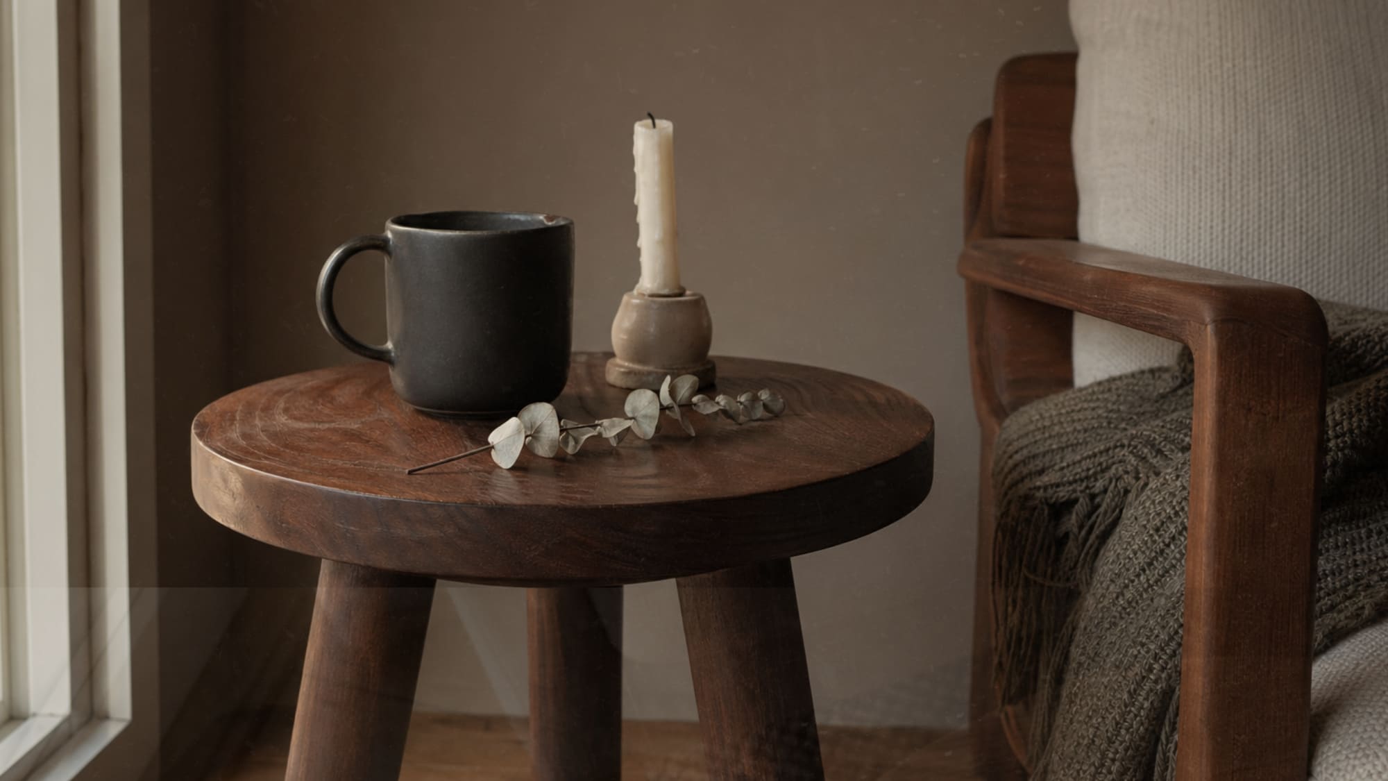 Low walnut three-legged side table in a Japandi reading nook styled with a matte ceramic mug, half-burned taper candle in clay holder, and dried eucalyptus stem on warm taupe wall