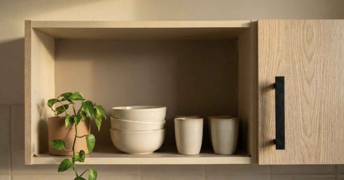 Rental kitchen upper cabinet with door removed and warm greige contact paper liner inside, styled with stoneware bowls, ceramic cups and a small pothos plant as open Japandi shelving