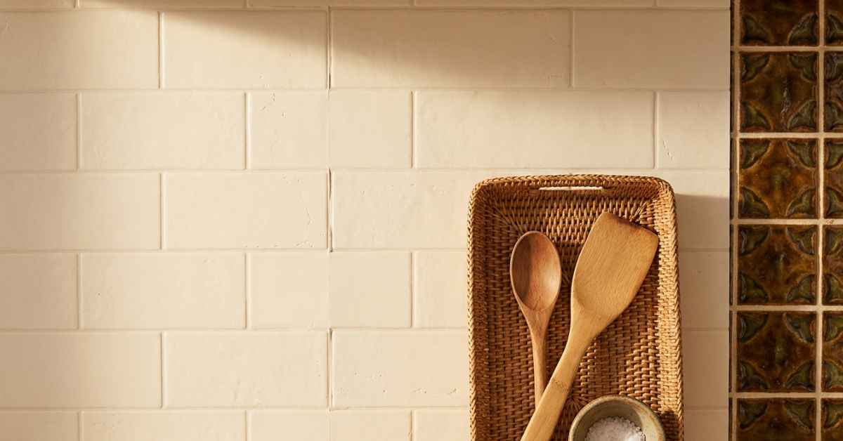 Rental kitchen backsplash with warm cream peel-and-stick subway tile panels applied over original dated tiles, rattan tray with wooden utensils leaning against the new surface