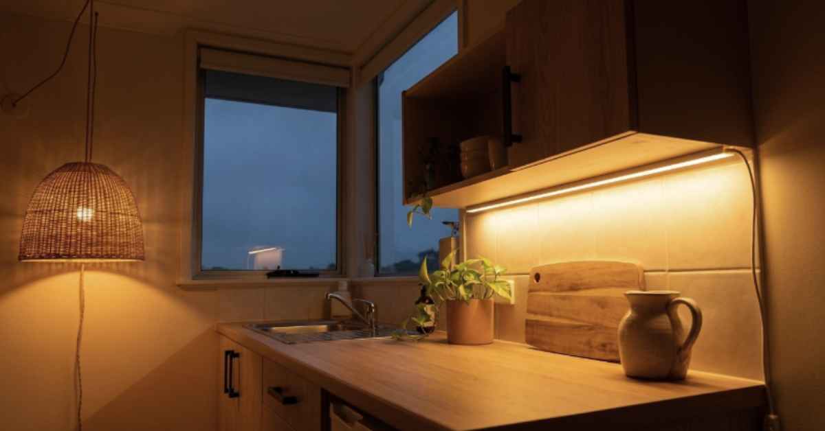 Small Japandi kitchen rental at dusk showing three warm light sources — under-shelf LED strip, rattan plug-in pendant and ambient lamp spill — with overhead light switched off